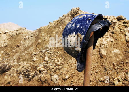 Blue safety hard hat on top of a spade at an archaeological excavation. Pile of dirt and stroke of blue sky in the background Stock Photo