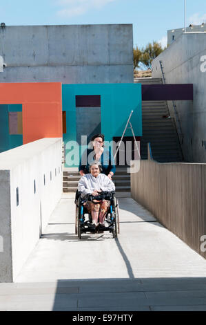 An elderly lady is pushed up a concrete disabled access ramp in a wheelchair by her carer or assistant on a sunny day. Stock Photo