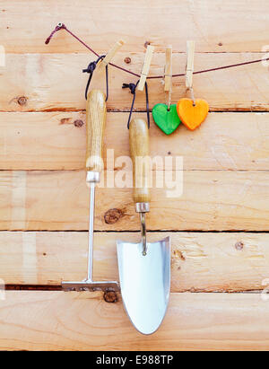 Garden implements hanging on wood with a small trowel and fork or rake alongside two colourful wooden hearts Stock Photo
