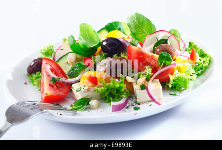 Healthy mixed Greek salad served on a white plate with silver fork containing crisp leafy greens, olives, feta, onion , tomato, Stock Photo