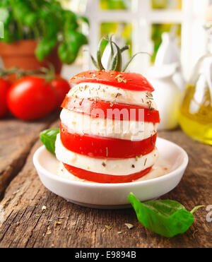 Preparing an individual gourmet cheese and tomato salad with a tower formed of alterning slices standing on a rustic wooden kitchen table Stock Photo