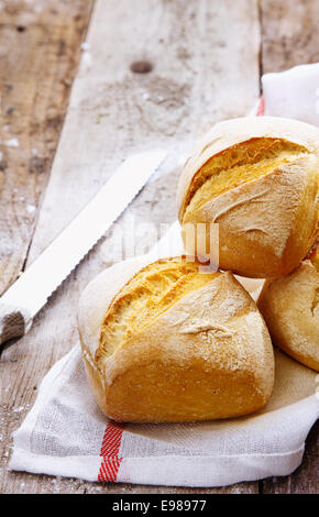 Close up of crusty golden bread rolls on a napkin with a bread knife on wooden boards Stock Photo