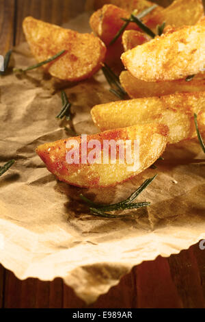 Golden baked or fried potato wedges with a spicy coating served as a takeaway on brown paper with fresh rosemary leaves Stock Photo