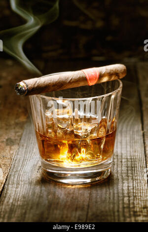 Burning Cuban cigar resting on glass of whisky on ice on an old wooden surface symbolic of masculinity Stock Photo