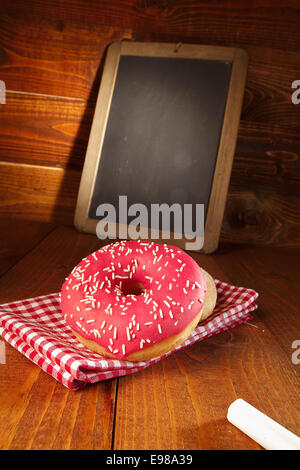 Freshly baked doughnut glazed with red icing and sprinkles served on a red and white checked cloth in front of a small blank chalkboard Stock Photo