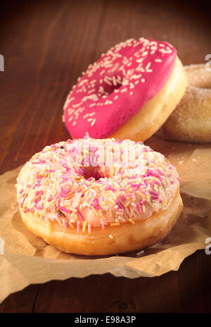 Two freshly baked golden doughnuts topped with colourful sprinkles on a grungy crumpled piece of brown paper against a wooden background with copyspace Stock Photo