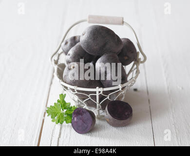 Fresh young whole lilac sweet potatoes in a wire basket with a sliced halved potato on the white wooden table in the foreground Stock Photo