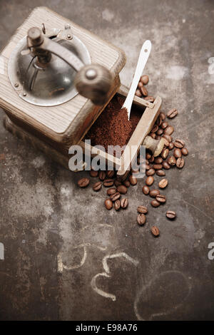 Overhead view of an old manual wooden coffee grinder with an open drawer filled with freshly ground coffee surrounded by scattered coffee beans on a grungy chalkboard with copyspace Stock Photo