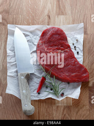 Portion of lean raw steak lying on a crumpled sheet of paper with a chefs knife and fresh rosemary with a red hot chilli pepper Stock Photo