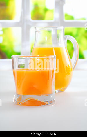 Tumbler of fresh healthy orange juice standing on a kitchen table alongside a jug full of juice with a sunny window and greenery behind Stock Photo