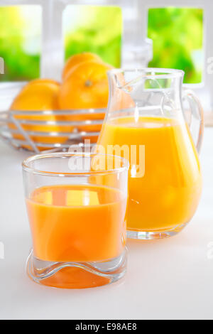 Freshly squeezed orange juice for breakfast standing on a white tabletop with a full glass tumbler and jug and a bowl of fresh oranges in the background against a sunny window with greenery Stock Photo