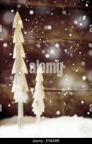 Two simple rustic Christmas trees cut out of wood standing in snow in front of a wooden wall with falling snowflakes for a festive Christmas winter background Stock Photo