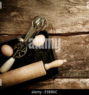 Baking concept with a wooden rolling pin and old egg beater resting on a baking tray with two fresh eggs on old wooden planking Stock Photo