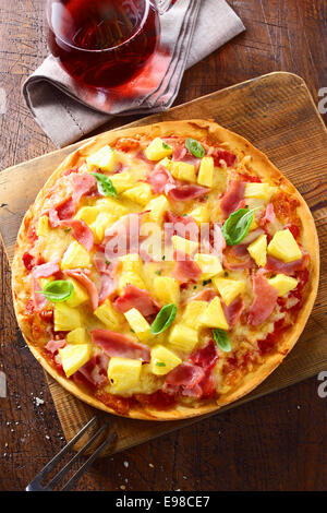 Overhead view of a delicious freshly baked ham and pineapple pizza on a thin crispy crust standing on an old wooden board on a kitchen table. More pizza at my port. Stock Photo