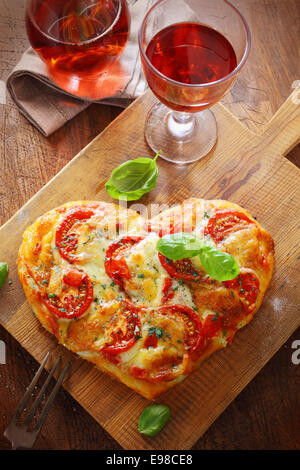 Overhead view of a tasty cheese and tomato vegetarian heart shaped pizza served on a wooden board with a glass of red wine