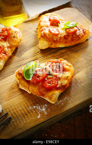 Small romantic heart shaped Italian pizzas with a freshly baked thick base topped with golden melted cheese and tomato and garnished with basil on a wooden board in the kitchen Stock Photo