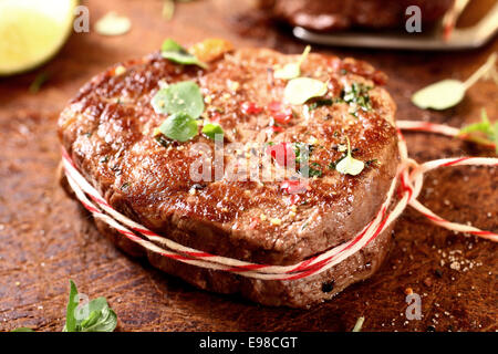 Thick juicy grilled lean beef steak medallions tied with colourful string and seasoned with chopped fresh herbs and spices on an old wooden board Stock Photo