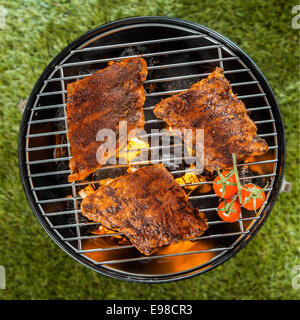 View from above of three delicious seasoned racks of rib grilling over a BBQ fire in a portable round metal barbecue on green grass Stock Photo