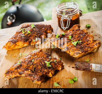 Seasoned ribs on a rustic wooden picnic table at a BBQ in the garden with fresh herbs and a glass jar of spicy marinade or basting sauce Stock Photo
