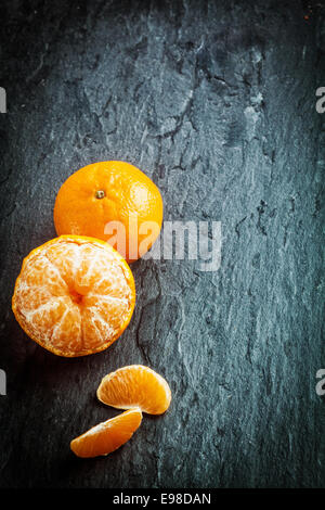 Peeled fresh clementine or tangerine with segments and a whole fruit arranged in a corner on a dark slate background with texture, copyspace and vignetting Stock Photo