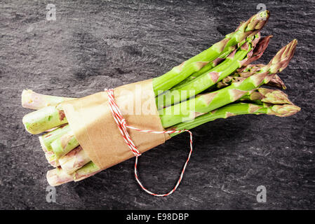Bundle of farm fresh green asparagus spears tied with brown paper and string on a dark textured slate background, overhead view Stock Photo