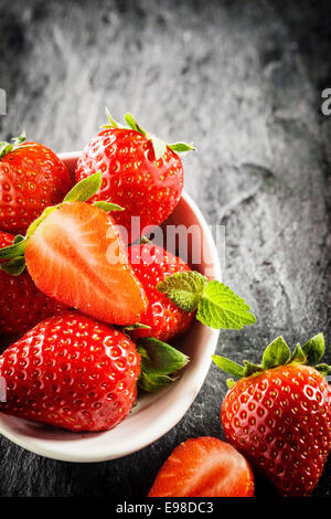 Bowl of ripe red strawberries with green stalks and one halved to show the juicy pulp, high angle view on a textured grey slate Stock Photo