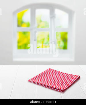 Red and white checked cloth neatly folded on top of an empty clean white table top under a window with a view of summer greenery in a country kitchen Stock Photo