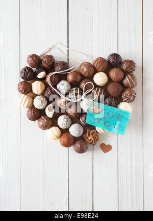 Heart shape made with various types of chocolate truffles and a blue tag with a romantic message over a white wooden table Stock Photo