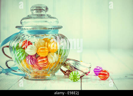 Low-angle view of an antique candy jar filled with colorful candies and ornate metal tongs on a wooden background