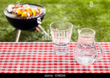 Glass of fresh water with a jug on a picnic table covered with a rustic red and white checkered cloth overlooking a green lawn and portable barbecue with food cooking Stock Photo