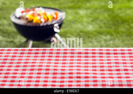 Empty picnic table background covered in a fresh country red and white checked cloth for your product placement or advertising with a barbecue on a green lawn behind Stock Photo