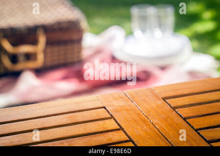 Empty slatted wooden picnic table for your product placement with a blurred wicker picnic hamper and rug with glasses and plates on a green lawn behind Stock Photo