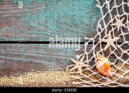 Rustic marine background with copyspace on weathered turquoise blue wooden boards decorated with diamond mesh fish net, starfish and a seashell on a bed of beach sand, mementos from a summer vacation Stock Photo