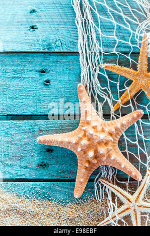 Colorful oceanic background with a marine still life of spiny starfish hanging on diamond mesh fish net on weathered rustic turquoise painted boards with copyspace and beach sand Stock Photo
