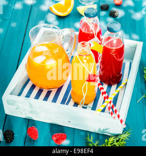 Fresh homemade orange and berry juice served in glass bottles and chilled with ice in a glass jug on a colorful, blue wooden picnic table on a wooden tray Stock Photo