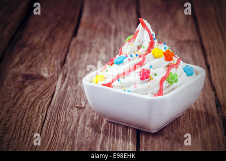 Ice cream party treat with a twirled bowl of vanilla ice cream topped with vibrant multicolored candy and sprinkles and drizzled with syrup on an old rustic wooden table Stock Photo