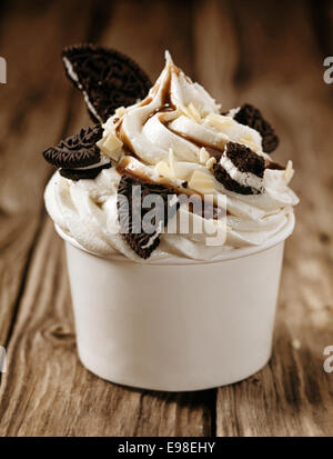 Takeaway tub filled with a swirl of vanilla ice cream with pieces of oreo biscuits and chocolate sauce, close up view on old wooden boards Stock Photo