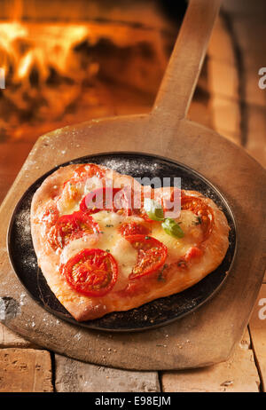 Romantic heart shaped Italian pizza on a platter and wooden board with the fire of a pizza oven visible behind Stock Photo