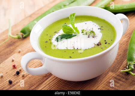 Close up of a bowl of green cream of pea soup garnished with cream and fresh herbs on a wooden surface with scattered pea pods Stock Photo