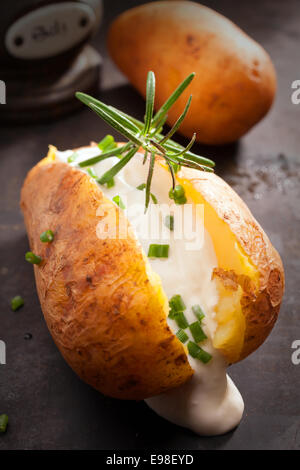 Sliced baked jacket potato cooked over a summer BBQ in foil with sour cream and chopped chives topped with rosemary for a delicious savory appetizer or vegetarian cuisine Stock Photo