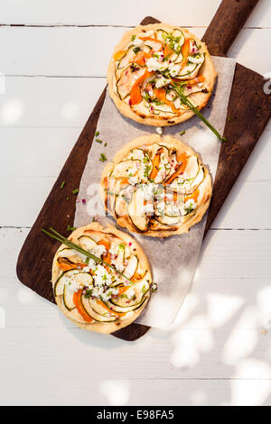 Three cooked delicious mini vegetarian pizzas on a long wooden board standing on a white painted wooden table outdoors in dappled sunlight, overhead view Stock Photo