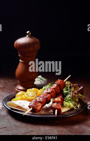 Two delicious spicy grilled pork kebabs served on a plate with fresh salad and tortilla on a rustic wooden table with a pepper mill and copyspace behind Stock Photo
