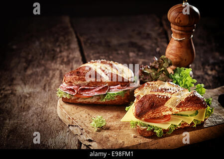 Crusty brown lye bread roll sandwiches with a cheese, salami, lettuce, tomato and cucumber filling on an old grunge board with a wooden pepper mill and copyspace Stock Photo