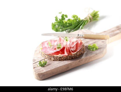 Preparing a delicious salami sandwich on crusty rye bread chopping a bunch of fresh parsley as a garnish, on an old wooden chopping board on white Stock Photo