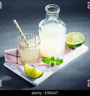 Bottle and glass of freshly prepared chilled lemon juice or lemonade with mint served with slices of fresh lime and lemon on a white board on a blue cloth with copyspace for a refreshing summer drink Stock Photo