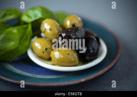 Cured green and black olives on a small plate with fresh basil leaves for use as ingredients in a salad or Italian cuisine Stock Photo
