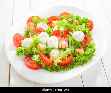 Fresh Italian salad with mozzarella cheese pearls on a bed of frilly lettuce with tomatoes and friend crunchy bread croutons served on a plate on white boards Stock Photo