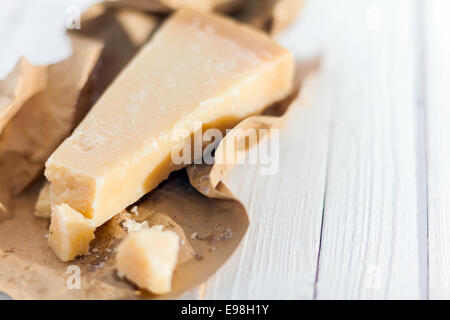 Wedge of hardy crumbly matured Italian Parmigiano-Reggiano cheese, or Parmesan, in a torn brown paper wrapper on a white wooden Stock Photo