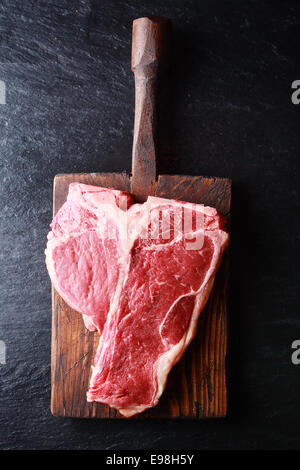 Overhead view of an uncooked t-bone steak on a rustic vintage wooden board on dark slate with copyspace Stock Photo