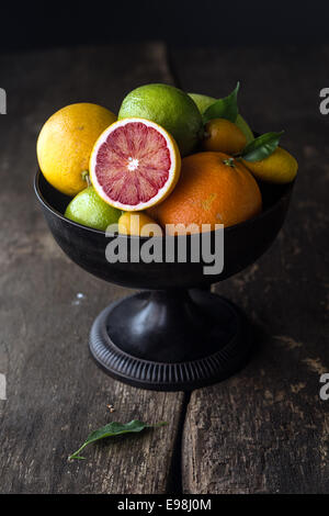 Pedestal bowl of assorted fresh citrus fruit with a halved blood orange displayed at the front on a rustic wooden background with shadows Stock Photo